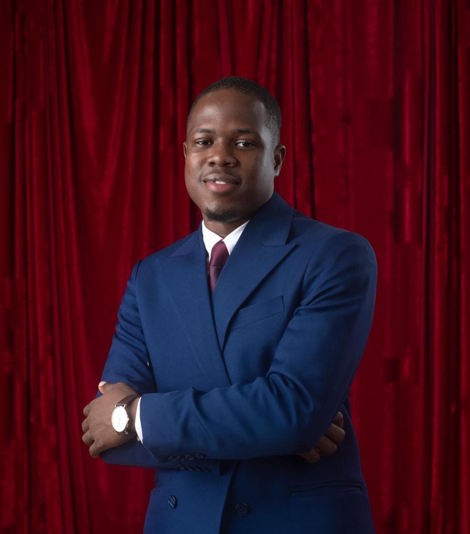 Adetola Adesote in a navy double-breasted suit and burgundy tie, arms folded, smiling, in front of a red velvet curtain